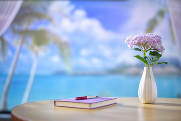 Seaside house interior wooden desk with notebook, pen, and a vase of purple flowers. Beautiful blurry view of beach landscape with blue sky and white clouds. Copy space.
