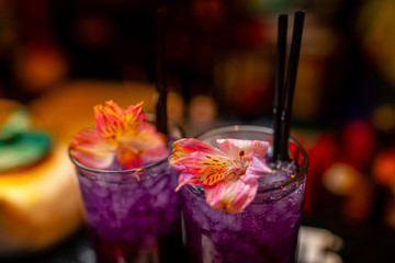 A refreshing violet alcoholic drink in the big cocktail glass decorated with a red flower on the bar counter. Close-up