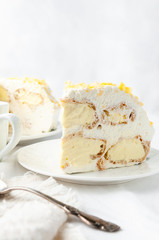 A piece of custard cake with whipped cream. Decorated with yellow chocolate chips. Home made. On a white plate. Near a spoon and a mug. In the background is a plate with cake and a white teapot. 