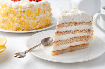 A piece of sponge cake with whipped cream and berries. garnished with pieces of white chocolate. Home made. On a white plate. In the background is a cake, a teapot and a white mug.
