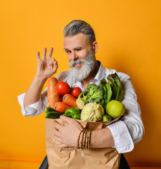 Adult grey haired stylish man with beard holding paper bag full of various healthy vegetarian goods and ingredients