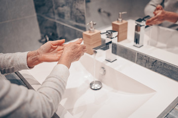 Fototapeta premium Hand washing lather liquid soap rubbing wrists handwash step senior woman rinsing in water at bathroom faucet sink. Wash hands for COVID-19 spreading prevention. Coronavirus pandemic outbreak.