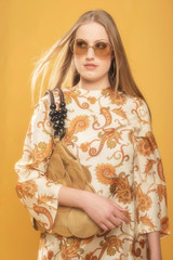 Fashionable blonde woman in retro paisley dress and sunglasses holds handbag.