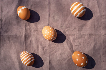 Painted 5 five brown eggs with various white patterns for Easter holiday on cotton purple tablecloth with sunlights and shadows. Top view. Eastern European Orthodox cultures. Easter concept