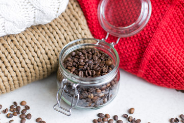 Macro shot of a transparent jar filled with coffee beans in the center of the photo, coffee beans around the jar and knitted sweaters in the background 