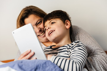 una mujer madre y su hijo con pluridiscapacidad (minusvalía, discapacidad) juegan con el ordenador portátil o tablet en la cama. van en pijama, y están alegres y sonrientes.