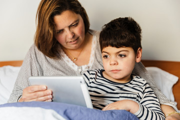 una mujer madre y su hijo con pluridiscapacidad (minusvalía, discapacidad) juegan con el ordenador portátil o tablet en la cama. van en pijama, y están alegres y sonrientes.