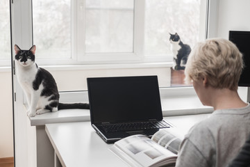 Girl sits at chair near table at home and leafs through a book with illustrations. Nearby sits black and white tuxedo cat. The concept of a break during work at home.