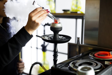 Fototapeta premium Women's hands hold hookah tongs and adjust the hot coals in a metal bowl. Black hookah stands in a restaurant or bar