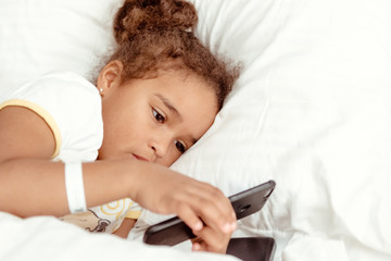 Cute little kid girl watching video on smartphone with smiley face alone on the bed, child using mobile phone with happy face at home. Stay at home quarantine coronavirus COVID-19 pandemic prevention.