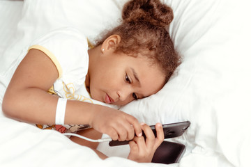 Obraz na płótnie Canvas Cute little kid girl watching video on smartphone with smiley face alone on the bed, child using mobile phone with happy face at home. Stay at home quarantine coronavirus COVID-19 pandemic prevention.