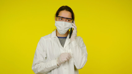 Young doctor in a medical mask, goggles and latex gloves is talking to patient on phone. Girl in a white coat on a yellow background holds a smartphone near her ear. Coronavirus, flu