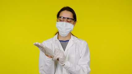 Young doctor in a medical mask and safety glasses puts latex gloves on her hands. Girl in a white coat on a yellow background