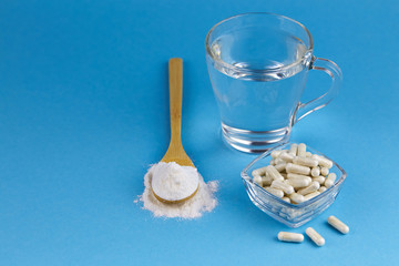 Collagen protein powder in spoon and collagen capsules in jar, a cup of water on blue  background. Natural beauty and health supplement for skin, bones, joints.