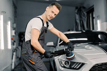 Fototapeta na wymiar Young handsome smiling Caucasian man, worker of auto detailing service, holds a polisher in the hand and polishes the car, looking and posing at camera. Car detailing and polishing concept