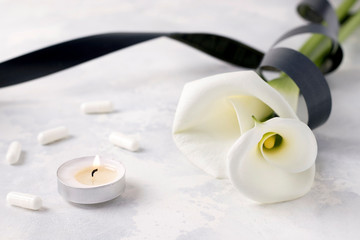 White calla flowers tied with a black mourning ribbon next to a burning candle with capsules and tablets on a white background. The concept of death due to the coronavirus pandemic