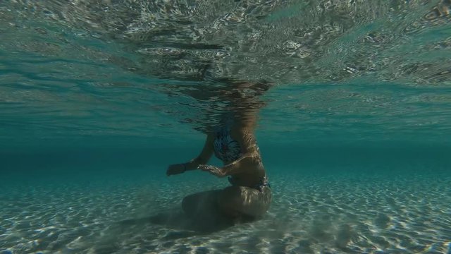 An underwater artistic shot of a girl sitting at the bottom of a white sandy beach, playing around the sand with there hands. Shot at sea level as the sunbeams pass through the water.