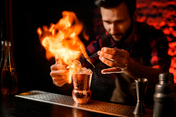 Fototapeta na wymiar bartender holds tweezers with spikelet over metal cup and makes fire flame.