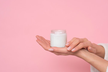 Commercial photo. Female hand holding a glass jar with cream on pink background. Moisturizing.