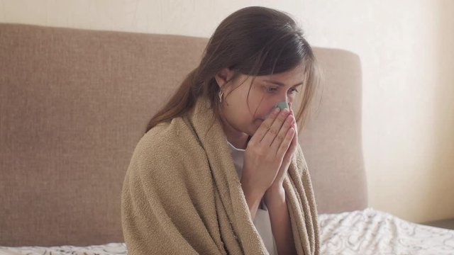 young woman trembling from cold wrapped in blanket sitting on the bed in bedroom interior, girl suffers from runny nose blowing in tissue, concept of illness, allergy and health