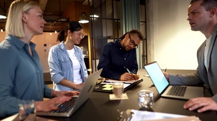Two diverse young businesspeople going over paperwork together while working with colleagues around an office table