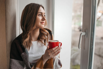 A young girl sits at the window with a cup of hot tea and a blanket, looks out the window at the street and smiles, quarantine coronovirus, isolation, stay home
