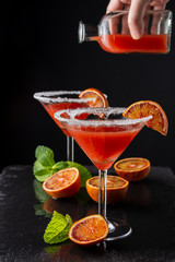 Close-up of two cocktail glasses with blood orange martini and hand with bottle and liquid, out of focus which is going to serve, in vertical