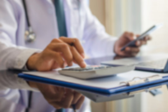 Blurred image of male doctor using mobile phone and calculator, work on laptop computer with clipboard on the desk at clinic or hospital. Medical healthcare costs ,fees and revenue concept
