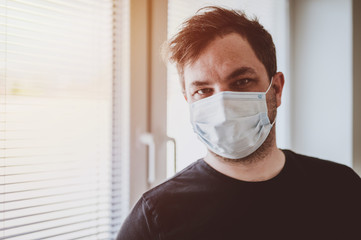 Man in quarantine during coronavirus COVID-19. Man with protective mask stands near window. Novel coronavirus 2019 COVID-19 theme. Quarantine during coronavirus pandemic