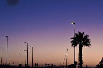 Schemenhafte Menschen als Schatten auf Strandpromenade bei lila Abendhimmel am Meer (Ibiza, Spanien)