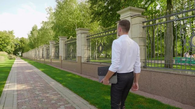 A businessman with a laptop in his hand walks through the park and looks around. A man freelancer in a white shirt and trousers goes to a meeting with a client. 4K footage
