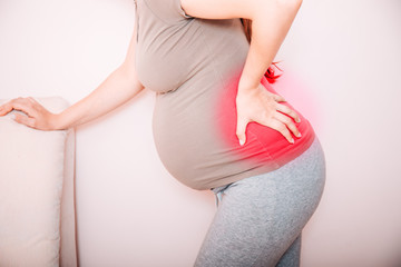Cropped view of young pregnant woman massaging her back. Horizontal shape, side view, copy space. Torso of pregnant woman with backache, pain in red.