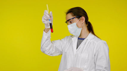 Doctor in a medical mask, goggles and latex gloves looks at the reaction in test tube. A young girl in a white coat on a yellow background holds hand a flask with red-black liquid