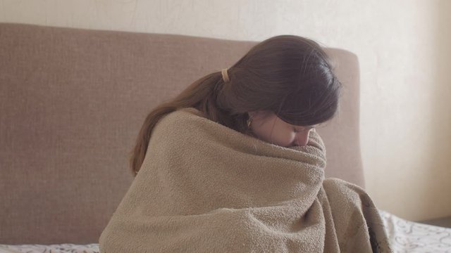 young woman trembling from cold wrapped in blanket sitting on the bed in bedroom interior, girl suffers from runny nose blowing in paper towel, concept of illness, allergy and health
