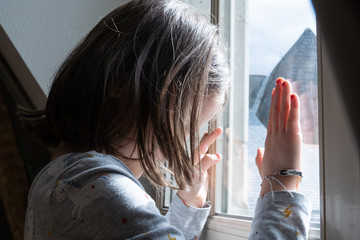 Little girl looking through the window with anxiety during coronavirus quarantine in the house. Stay home concept for families with children. Psychological problems in children in isolation. - obrazy, fototapety, plakaty