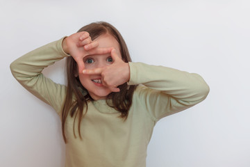 A little girl in a light sweater on a white gray background depicts the process of photography with her hands