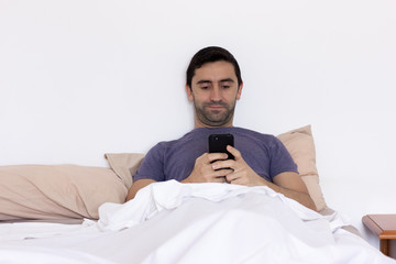 Young man lying on bed pillow writing message on cellphone in bright bedroom. Scruffy looking guy...
