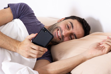 Smiling man lying in bed talking on cellphone in bright morning. Person starting day looking at smart phone in bedroom. Mobile addiction, connection, good news concepts