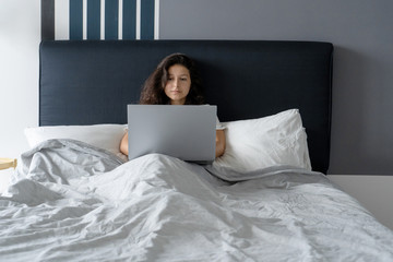Beautiful brunette girl in bed with a laptop. Work from morning to late night. Electronic gadgets before bedtime or in the morning.