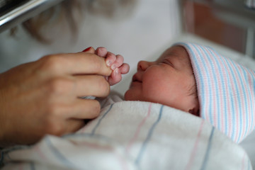 A mother holds her newborn infants hand in the hospital bassinet 