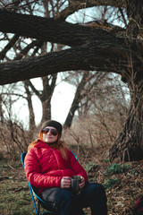 The girl is dressed in a red down jacket holding metal cup. Sunglasses and hat. Sits in a folding camping chair. Light weight and compact design. Outdoor camp. Nature. Vertical photo