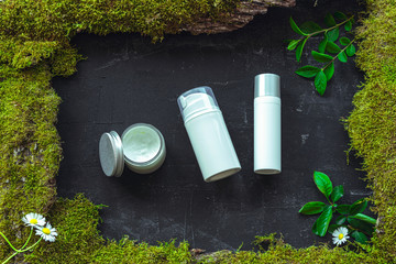Moisturizing cream jar, serum and skin tonic in white unbranded blank packages on black background with moss and greenery. Natural cosmetics concept. Top view, flat lay.