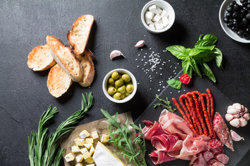 Tapas concept. Prosciutto, sausage chorizo, olives, baguette, arugula, basil and brie cheese on a black slate board. Antipasti concept.