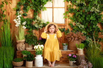 A little girl sits on porch of wooden house, around green houseplants and flowers. Kid is in yellow dress, straw hat. Concept of childhood. Gardening. Rustic terrace, porch. Child playing in backyard