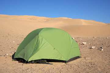 The light green tent and small stones around it in the desert near the big sandy hill, the blue sky.