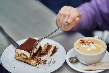 Girl hand with a fork breaks off a piece of cheesecake, a cup of coffee is on the table next. Close-up.
