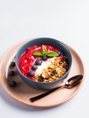 Yogurt smoothie bowl with blended raspberries, blueberries and oat matcha granola on pastel background. Delicious healthy breakfast concept on gray blue
