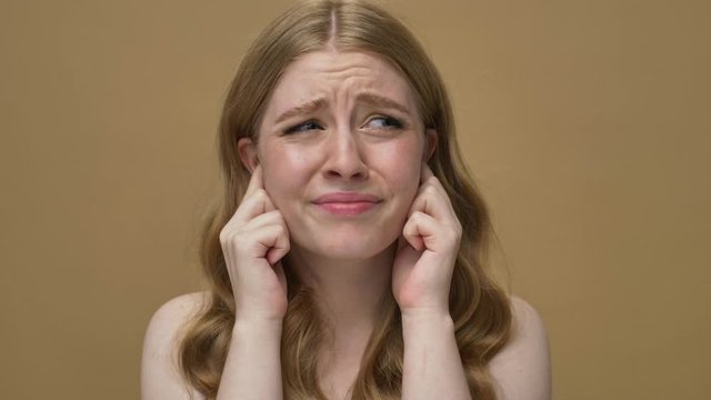 A displeased young half-naked woman with long hair is covering her ears with her fingers isolated over beige background