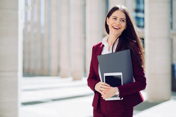 Portrait of business woman dressed in maroon suit, holds tablet and folder, smiling widely outdoor. Business people concept.