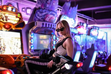 A beautiful sexy blonde girl in dark glasses poses on a motorcycle racing simulator under neon lights. Girl under the neon lights. Video games, virtual reality, slot machines. Girl gamer.